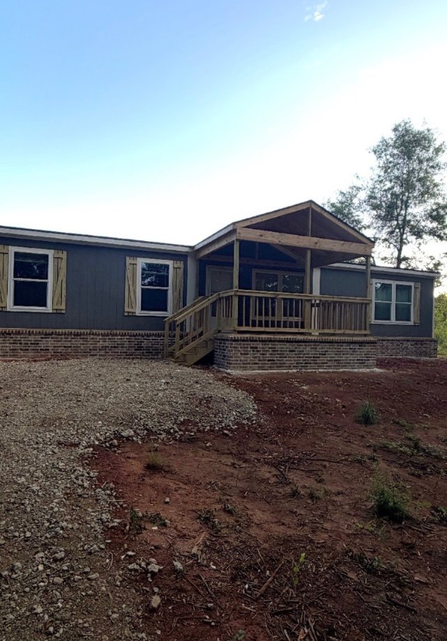 Covered wood porch with railings, side steps and a brick base in front of gray siding.